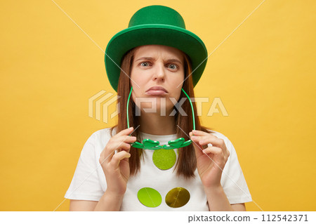 Irish holiday boring party. Sad upset unhappy woman wearing green hat holding clover glasses standing isolated over yellow background looking at camera with unhappy disappointed expression Irish holiday boring party. Sad upset unhappy woman wearing green hat holding clover glasses standing isolated over yellow background looking at camera with unhappy disappointed expression 112542371