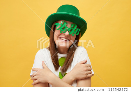 Festive celebration St. Patrick's day. Pleased pretty woman wearing green hat and clover glasses standing isolated over yellow background hugging herself smiling happily Festive celebration St. Patrick's day. Pleased pretty woman wearing green hat and clover glasses standing isolated over yellow background hugging herself smiling happily 112542372