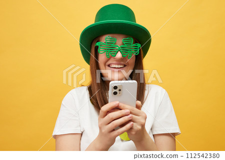 Smiling cheerful woman wearing green hat and clover glasses standing isolated over yellow background scrolling online with mobile phone browsing web pages 112542380
