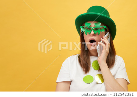 Shocked astonished woman wearing green hat and clover glasses standing isolated over yellow background talking on smartphone copy space for advertisement Shocked astonished woman wearing green hat and clover glasses standing isolated over yellow background talking on smartphone copy space for advertisement 112542381