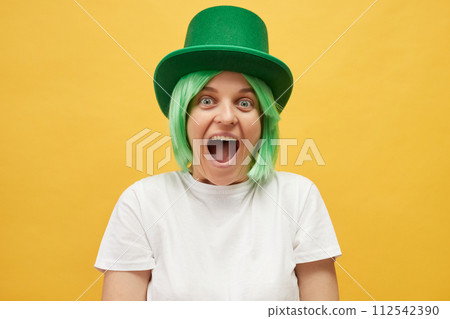 Overjoyed excited amazed beautiful woman with green hair wearing leprechaun hat standing isolated over yellow background screaming wow OMG with happy face 112542390