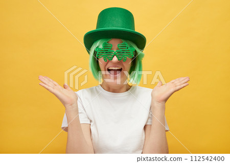 Funny woman with green hair wearing leprechaun hat and shamrock glasses posing isolated over yellow background spreading her face exclaiming with excitement Funny woman with green hair wearing leprechaun hat and shamrock glasses posing isolated over yellow background spreading her face exclaiming with excitement 112542400