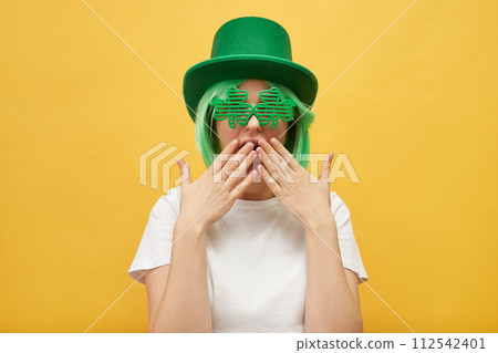 Astonished surprised woman with green hair wearing leprechaun hat and shamrock glasses posing isolated over yellow background looking with shocked eyes Astonished surprised woman with green hair wearing leprechaun hat and shamrock glasses posing isolated over yellow background looking with shocked eyes 112542401