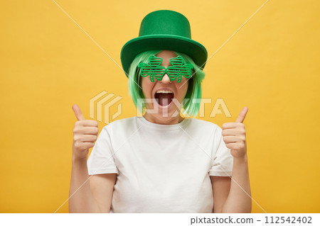 Overjoyed excited woman with green hair wearing leprechaun hat and shamrock glasses posing isolated over yellow background showing like gesture thumbs up Overjoyed excited woman with green hair wearing leprechaun hat and shamrock glasses posing isolated over yellow background showing like gesture thumbs up 112542402