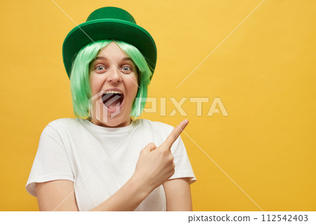 Amazed excited woman with green hair wearing leprechaun hat posing isolated over yellow background pointing at copy space for promotional text indicating at advertisement area Amazed excited woman with green hair wearing leprechaun hat posing isolated over yellow background pointing at copy space for promotional text indicating at advertisement area 112542403