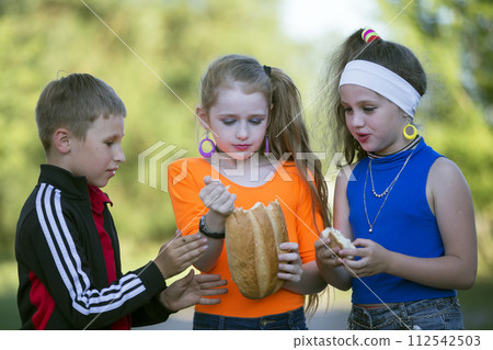 Cheerful children, a boy and two girls in bright clothes are eating a roll on the street. Cheerful children, a boy and two girls in bright clothes are eating a roll on the street. 112542503