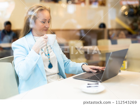 Business lunch - businesswoman sits at table in cafe by window with a laptop and drinks coffee. View through window 112543269