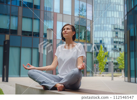 De-stress, unwind idea. peaceful female freelance employee practicing yoga exercises while sitting in bunch under glass skyscrapers after hard working day in office, holding hands in mudra 112546547