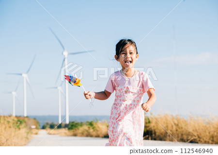 A joyful child holding pinwheels runs near windmills embodying wind energy education. Clean electricity and sustainable industry visuals in a picturesque wind turbine landscape. 112546940
