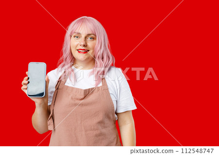Great Mobile Offer. Excited happy 30s woman with pink hair in apron holding Smartphone in her hand, Emotionally Reacting To New App. Female posing on red background, studio portrait. 112548747