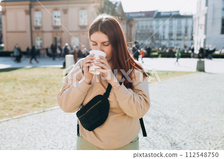 Happy young caucasian woman in good mood walks with coffee around city during day. Lifestyle concept. 30s girl holding takeaway coffee cup on urban background 112548750