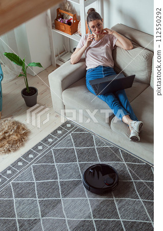 Robotic vacuum cleaner cleaning carpet, woman using laptop sitting on sofa at home Robotic vacuum cleaner cleaning carpet, woman using laptop sitting on sofa at home 112550292
