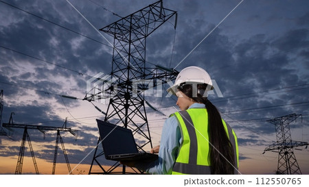 Woman electrician marks notes online on laptop observing power transmission lines at twilight countryside closeup. Engineer tests system with laptop at power generation plant under dramatic sky Woman electrician marks notes online on laptop observing power transmission lines at twilight countryside closeup. Engineer tests system with laptop at power generation plant under dramatic sky 112550765