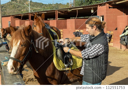 Portrait of confident adult horseman adjusting saddle of horse before horseback ride in country club 112551753
