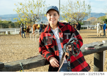 Smiling European young woman instructor holding horse halter or reins during working day at horse club 112551764