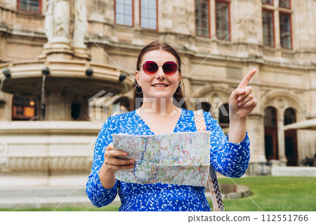 Young female tourist with paper map pointing finger outdoors. Urban lifestyle concept, summer time. Vienna State Opera. Concept of travel, tourism and vacation in city. 112551766
