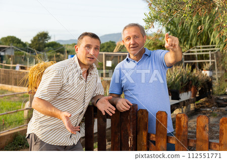 Two male farmers talking outside next to wooden fence on background with farm Two male farmers talking outside next to wooden fence on background with farm 112551771