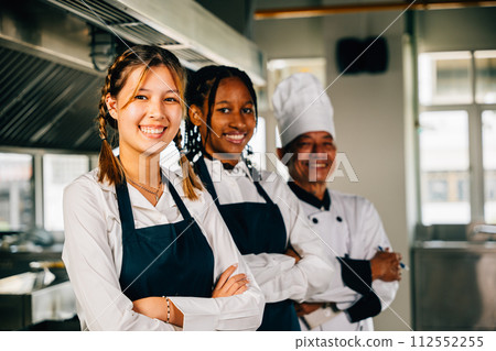 In a professional kitchen smiling chefs in uniform pose for a portrait. Displaying expertise togetherness and teamwork. Teaching colleagues working together. In a professional kitchen smiling chefs in uniform pose for a portrait. Displaying expertise togetherness and teamwork. Teaching colleagues working together. 112552255