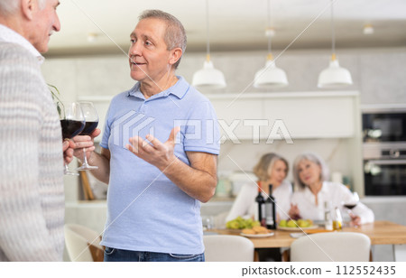 Elderly men have withdrawn from their female companions, stand with glass of wine in hands, chatting 112552435