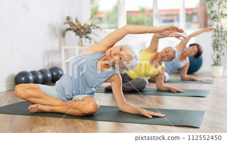 Old woman practicing lotus pose of yoga with side bend in training room Old woman practicing lotus pose of yoga with side bend in training room 112552450