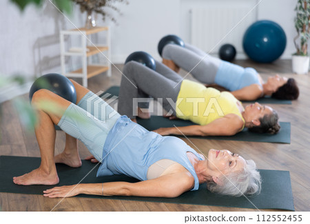 Old woman practicing bridge pose with softball between legs during fitness classes 112552455