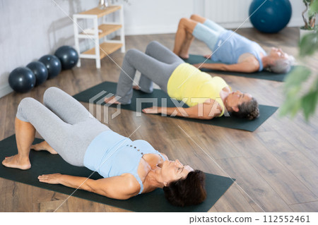 Elderly woman in company of hatha yoga lovers performs bridge pose, setu bandha sarvangasana 112552461