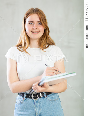 Portrait of cheerful teen girl in casual clothes writing in spiral notepad against concrete gray wall. Free space for mock up Portrait of cheerful teen girl in casual clothes writing in spiral notepad against concrete gray wall. Free space for mock up 112552998