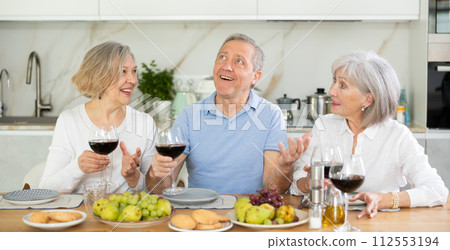 Mature married couple with female friend are sitting at table, enjoying food and wine, chatting. 112553194