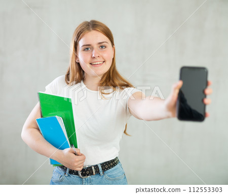 Smiling teenage girl, student with notebooks under her arm and mobile phone with blank black screen at gray wall background. Free copy space for advertisement 112553303