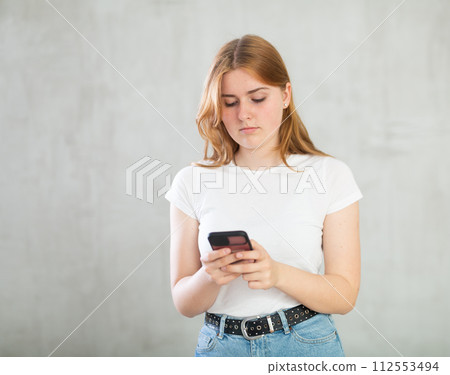 Positive teenage girl, student in casual clothes surfing Internet on smartphone against gray wall background Positive teenage girl, student in casual clothes surfing Internet on smartphone against gray wall background 112553494