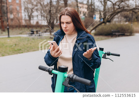 Portrait of young woman with confused face, holding smartphone near scooter, using mobile application on urban background Portrait of young woman with confused face, holding smartphone near scooter, using mobile application on urban background 112553615