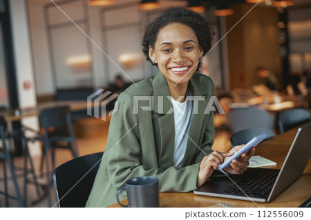 A woman in a suit smiles at her laptop and phone on the table A woman in a suit smiles at her laptop and phone on the table 112556009