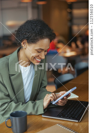 A woman sits at a table with a laptop and tablet, smiling in her blazer 112556010