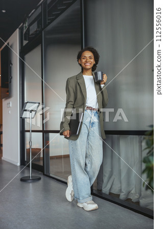 A woman in formal wear holds a coffee cup and laptop in a hallway A woman in formal wear holds a coffee cup and laptop in a hallway 112556016
