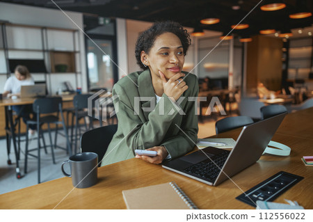 A woman is seated at a table with a laptop and a cell phone in nice modern coworking office 112556022