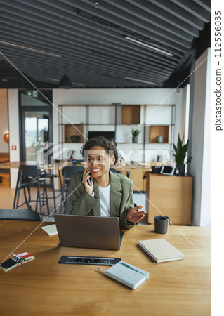 A woman seated at a desk with a laptop, talking on a cell phone in nice modern coworking office A woman seated at a desk with a laptop, talking on a cell phone in nice modern coworking office 112556035