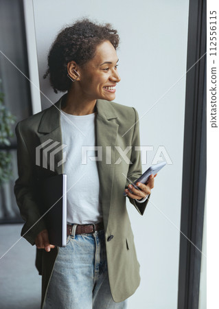 Woman in formal wear, leaning against wall with laptop and cell phone, smiling 112556115