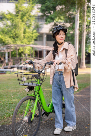 A young Asian female college student wearing a bike helmet is pushing her bicycle in a green park. A young Asian female college student wearing a bike helmet is pushing her bicycle in a green park. 112558348