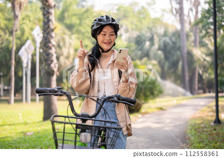 A positive Asian woman is checking a map on her smartphone while riding a bike in a green park. A positive Asian woman is checking a map on her smartphone while riding a bike in a green park. 112558361