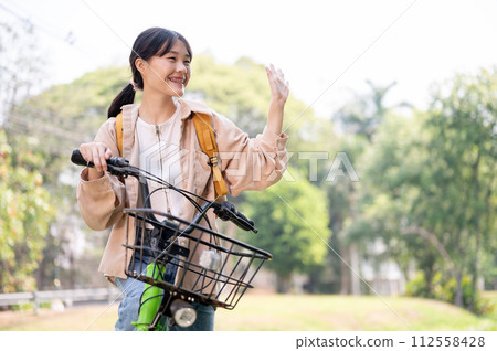 A cheerful Asian female college student waves her hand, greeting her friends while riding her bike. 112558428