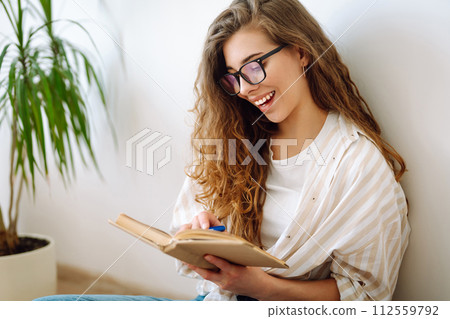 Young woman reading book, writing notes, doing homework at home. Concept of rest, relaxation. Young woman reading book, writing notes, doing homework at home. Concept of rest, relaxation. 112559792