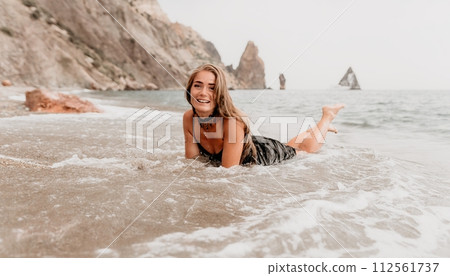 Woman summer travel sea. Happy tourist in black dress enjoy taking picture outdoors for memories. Woman traveler posing on sea beach surrounded by volcanic mountains, sharing travel adventure journey 112561737