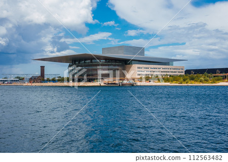 A modern, layered architectural marvel reflects the sky on its glass and metal facade, likely the Copenhagen Opera House, set against a calm waterfront under a partly cloudy sky. 112563482