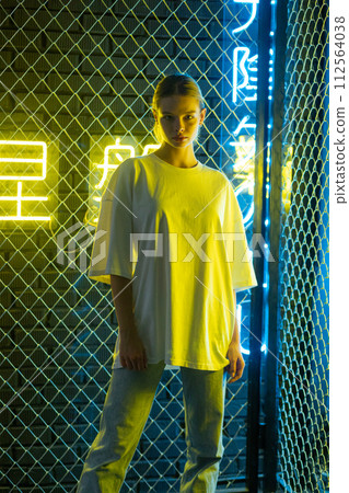 A beautiful blonde girl in a white oversized T-shirt and blue jeans posing against the background of neon hieroglyphs 112564038
