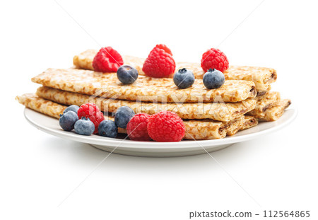 A white plate is arranged with pancakes covered in a variety of berries and blueberries isolated on white background. 112564865