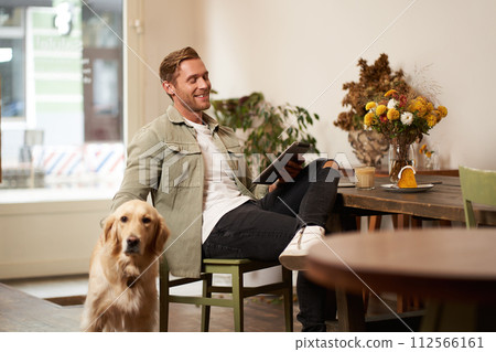Portrait of handsome young man sits in cafe with his dog, petting golden retriever, reading news on tablet, relaxing with cup of coffee 112566161