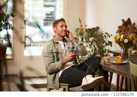 Portrait of young handsome man in casual clothes, sits in cafe and enjoys favourite song, listens to music in wireless headphones, vibing to the relaxing tunes in earphones, holding smartphone 112566170