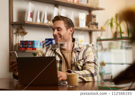 Lifestyle portrait of young businessman, freelancer sitting in cafe, drinking coffee and using laptop, working on online project, digital nomad doing his task, has remote job, looking satisfied Lifestyle portrait of young businessman, freelancer sitting in cafe, drinking coffee and using laptop, working on online project, digital nomad doing his task, has remote job, looking satisfied 112566193