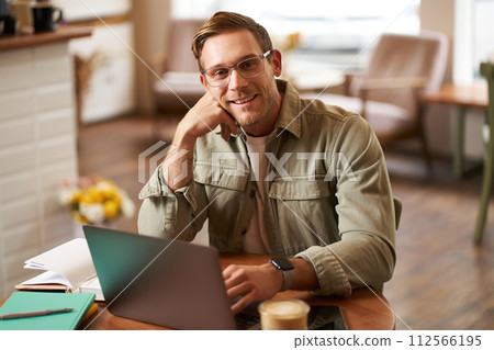 Remote work and freelance concept. Handsome man in glasses sits with notebook and laptop, studying online, working remotely from coffee shop, sitting in cafe with computer Remote work and freelance concept. Handsome man in glasses sits with notebook and laptop, studying online, working remotely from coffee shop, sitting in cafe with computer 112566195