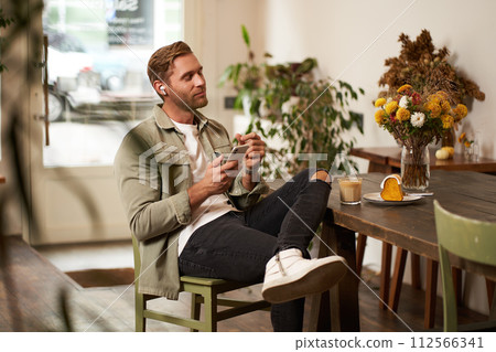 Portrait of young handsome man in casual clothes, sits in cafe and enjoys favourite song, listens to music in wireless headphones, vibing to the relaxing tunes in earphones, holding smartphone Portrait of young handsome man in casual clothes, sits in cafe and enjoys favourite song, listens to music in wireless headphones, vibing to the relaxing tunes in earphones, holding smartphone 112566341